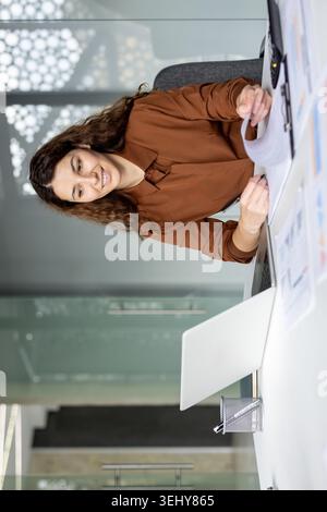 Donna d'affari ispanica sorridente in una camicia marrone seduta alla scrivania dell'ufficio, che lavora con un computer portatile e rivede i documenti finanziari in una clipboard. Foto Stock