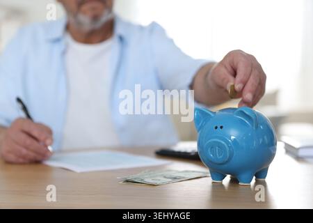 Fondo pensione. Uomo anziano che mette soldi nel salvadanaio al tavolo al chiuso, attenzione selettiva Foto Stock