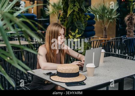 La donna usa il computer portatile mentre siede al tavolo sulla terrazza verde del ristorante. Lavoratrice in telelavoro, area di lavoro all'aperto con notebook, donna Foto Stock