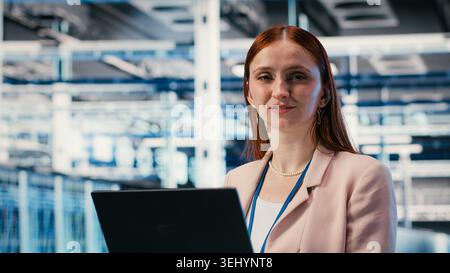 Donna felice in sala server che utilizza un notebook per migliorare l'efficacia operativa dell'infrastruttura. Gioiosa ingegnere donna nel data center che utilizza un notebook per monitorare le metriche di RIGS, telecamera B. Foto Stock