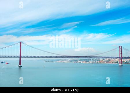 Un grande ponte sospeso rosso, il 25 aprile, si estende attraverso un ampio fiume con navi in acqua e uno skyline della città Foto Stock