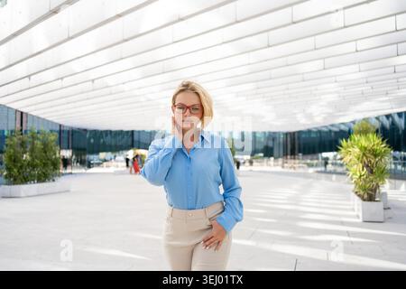 Donna d'affari bionda che indossa occhiali che cammina con sicurezza all'entrata del moderno edificio di uffici con facciata in vetro e spazio aperto Foto Stock