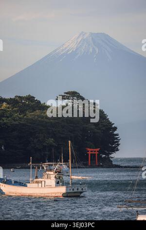 Numazu, Giappone - 4 maggio 2026: Vista del Monte Fuji che torreggia sulla spiaggia di Mihama con un cancello torii rosso annidato tra gli alberi e una barca da pesca in primo piano Foto Stock