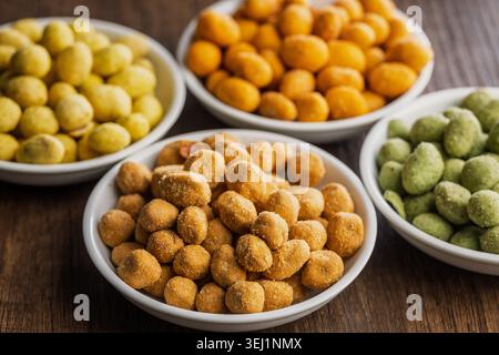 Arachidi rivestite assortite servite in piccole ciotole bianche su un tavolo di legno, caratterizzate da croccanti e aromatizzati in giallo, arancione e verde. Ciotole colorate ideali per feste, antipasti o spuntini informali. Foto Stock