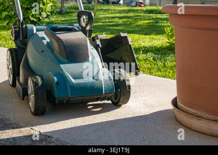 Un rasaerba elettrico blu parcheggiato su un sentiero di cemento accanto a un grande vaso di terracotta. Sullo sfondo è visibile erba verde rigogliosa, a indicare un pozzetto-m. Foto Stock