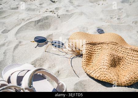 Occhiali da sole, un cappello di paglia e sandali bianchi sono posizionati sulla spiaggia di sabbia. La scena cattura un'atmosfera di vacanza estiva rilassata. Foto Stock