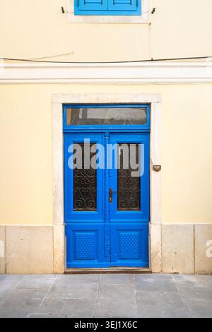 Pirano, Slovenia. 26 maggio 2026. Una doppia porta blu brillante con inserti in metallo ornato è inserita in una parete color crema. Sopra la porta, un piccolo windo blu Foto Stock