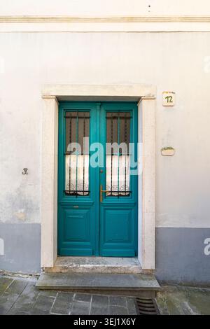 Pirano, Slovenia. 26 maggio 2026. Una luminosa porta doppia color ottanio si trova all'interno di un telaio in pietra su un edificio bianco. Un numero civico 12 è esposto sulla parete Foto Stock
