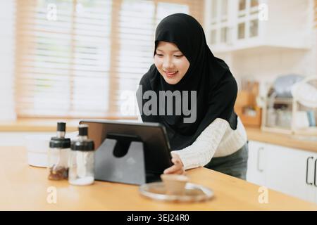 La giovane donna musulmana araba in hijab lavora con un tablet nella cucina domestica sorridendo felice. Foto Stock