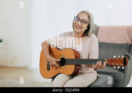 Sana anziana donna asiatica musicista che suona la chitarra amante della musica nel salotto di casa. Foto Stock
