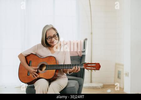 Sana anziana donna asiatica musicista che suona la chitarra amante della musica nel salotto di casa. Foto Stock