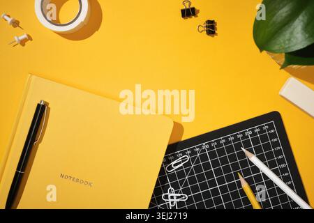 Notebook giallo con penna nera su scrivania gialla brillante. Tappetino da taglio nero, matite, graffette, graffette per rilegatura, rotolo di nastro, spinotti e piante. Copyspace Foto Stock