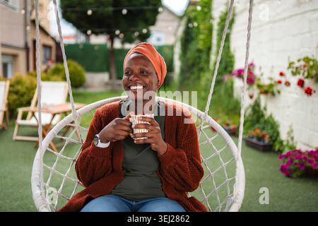Giovane donna nera che si rilassa nell'altalena del cortile tenendo il caffè Foto Stock