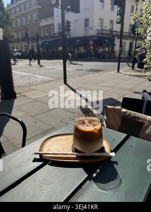 Un bicchiere di rinfrescante caffè ghiacciato in piedi su un piccolo vassoio di legno da portata su un tavolo da terrazza verde scuro in un caffè di strada a Belgravia, Londra. Foto Stock