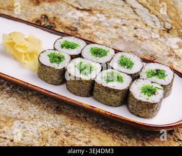 Cibo, panini di sushi ripieni di vibranti ingredienti verdi riposano su un lungo piatto di legno. Le fette di zenzero sono posizionate accanto ai panini, offrendo una freschezza di Foto Stock