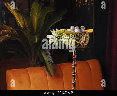 Cibo, esotico narghilè servito in una ciotola di ananas intagliata con kiwi e fette di arancia, presentato su un alto piedistallo in un elegante interno lounge con caldo mattino Foto Stock