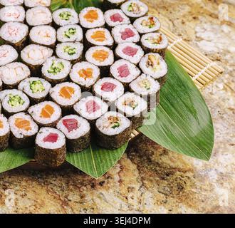 Cibo, Una varietà di coloratissimi sushi roll seduti su un piatto di bambù, con diverse farciture e condimenti. Il piatto poggia su una superficie in pietra, inv Foto Stock