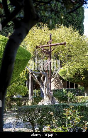 Vieste - Puglia - Italia - 08-29-2025: La scultura del crocifisso in metallo arrugginito sorge in un giardino curato, incorniciato da alberi e siepi, con un edificio in pietra a Foto Stock
