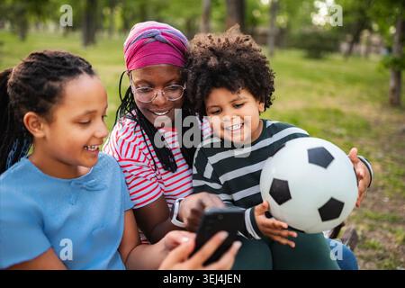 Madre e bambini guardano lo smartphone mentre si rilassano nel parco Foto Stock
