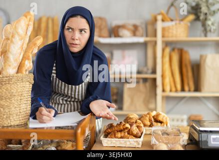 Donna europea in hijab che esamina i documenti mentre gestisce la panetteria Foto Stock