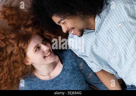 Felice giovane coppia multirazziale sdraiata insieme, sorridente e che si guarda l'un l'altra Foto Stock
