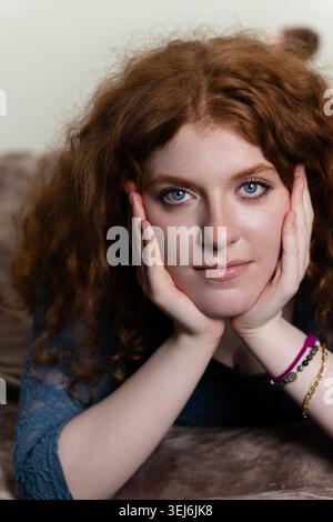 Giovane donna con capelli ricci rossi che riposa la testa sulle mani, guardando direttamente Foto Stock