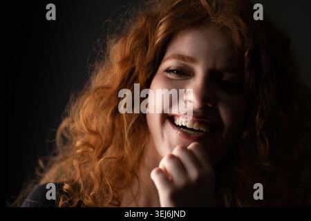 Giovane donna con capelli ricci rossi che ride con gli occhi chiusi su uno sfondo scuro Foto Stock