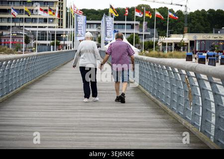 Groemitz - Schleswig-Holstein - Germania - 06-28-2025: Una coppia di anziani cammina mano nella mano su una passerella di legno con ringhiere metalliche, bandiere e mari Foto Stock