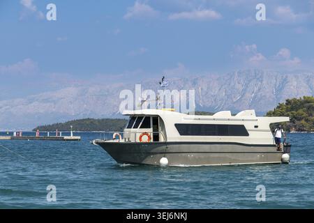 Nydri - Isole Ionie - Grecia - 08-07-2025: Una nave passeggeri con bandiera greca si muove attraverso l'acqua vicino al molo, con montagne e una parte di clo Foto Stock