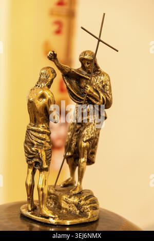 Sessa Aurunca - Campania - Italia - 08-29-2025: Piccola scultura religiosa dorata di una scena di battesimo con due figure e un bastone a croce, esposta in indoo Foto Stock
