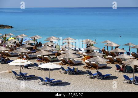 Nydri - Isole Ionie - Grecia - 08-09-2025: File di ombrelloni in paglia e tessuto con lettini da mare, spiaggia sabbiosa accanto al mare turchese, dove le persone Foto Stock