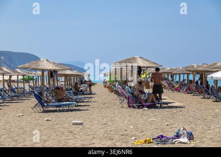 Nydri - Isole Ionie - Grecia - 08-09-2025: Gli amanti della spiaggia si rilassano sui lettini sotto gli ombrelloni di paglia sulla spiaggia sabbiosa, con mare turchese nel backgroun Foto Stock