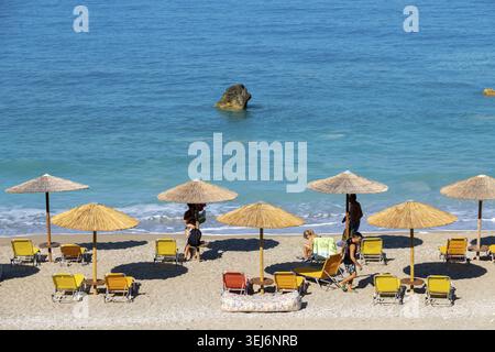 Nydri - Isole Ionie - Grecia - 08-09-2025: Spiaggia con ombrelloni di paglia, lettini gialli e arancioni, persone che si rilassano sulla spiaggia sabbiosa e tur Foto Stock