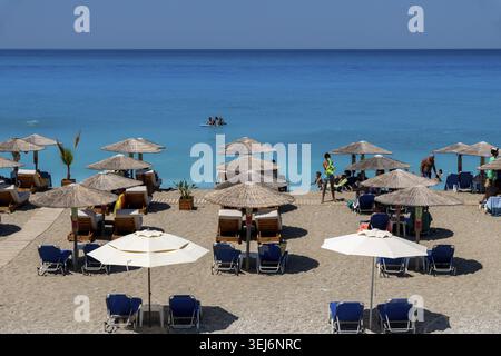 Nydri - Isole Ionie - Grecia - 08-09-2025: File di ombrelloni di paglia e lettini da sole fiancheggiano la spiaggia sabbiosa, con persone che si rilassano e nuotano nella Foto Stock