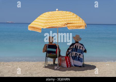 Nydri - Isole Ionie - Grecia - 08-09-2025: Due persone sedute sotto un ombrello arancione a motivi geometrici su una spiaggia sabbiosa, di fronte al mare turchese calmo con la nave Foto Stock