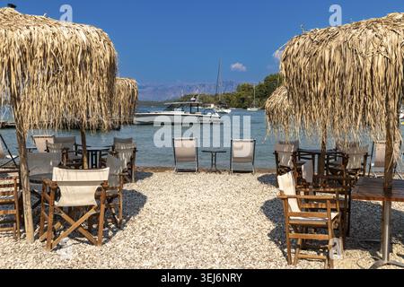 Nydri - Isole Ionie - Grecia - 08-07-2025: Sedie in legno e ombrelloni di paglia su una spiaggia di ciottoli con barche ancorate in acque calme blu vicino alla riva Foto Stock