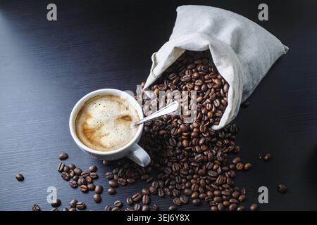 Tazza di caffè espresso e chicchi di caffè sparsi da una borsa di tela su sfondo nero in legno Foto Stock