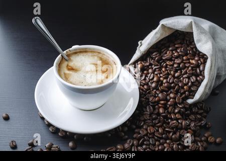 Tazza da caffè, piattino e chicchi di caffè sparsi da una borsa di tela su un tavolo di legno nero Foto Stock