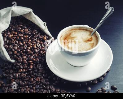 Ottimo caffè appena fatto. Foto della tazza di caffè e dei chicchi di caffè sparsi da una borsa di tela Foto Stock