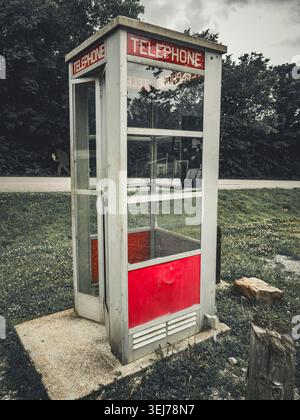 Cabina telefonica esterna vintage in vetro e metallo in piedi su una strada erbosa sotto un cielo coperto con sfondo boschivo Foto Stock