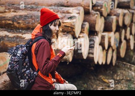 Donna siede sui registri nella foresta mentre utilizza lo smartphone. Moderno stile di vita nomade, hobby all'aperto per donne, invecchiamento attivo e benessere. Foto Stock