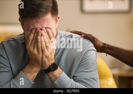 Primo piano di un giovane uomo adulto caucasico che copre il viso, mentre un amico maschio lo conforta con la mano sulla spalla, esprimendo sostegno emotivo. Ideale per la salute mentale, l'amicizia, la consulenza Foto Stock