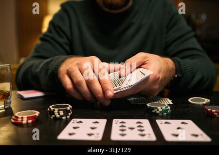 Giovane adulto che tiene in mano carte da gioco e gioca con le patatine da poker al tavolo, mostrando rischio e strategia. Utile per marketing di casinò, contenuti di scommesse, campagne di sensibilizzazione sulle dipendenze Foto Stock