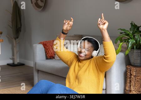Donna afroamericana che canta e balla nel salotto con cuffie e maglione senape Foto Stock
