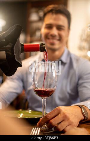 Primo piano di vino rosso versato in un bicchiere durante una cena romantica in un ristorante accogliente. Giovane sorridente sullo sfondo mentre si gode un el Foto Stock