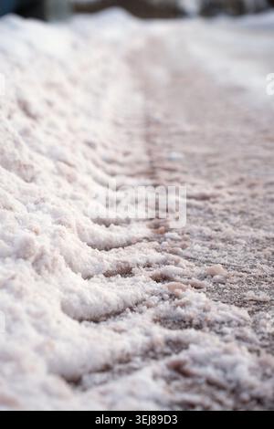 vista ravvicinata delle tracce degli pneumatici pressate nella neve soffusa lungo la strada in una luce invernale soffusa, rivelando una consistenza di colore bianco-rossastro e marrone tenue Foto Stock