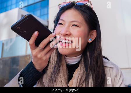Giovane donna asiatica che tiene il telefono che invia una nota vocale e sorride fuori dall'area urbana Foto Stock