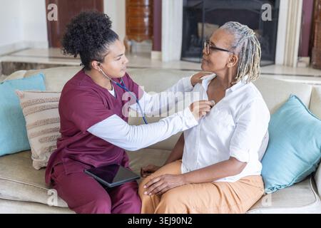 Infermiera in scrub usando lo stetoscopio su una donna afroamericana anziana sul divano con tablet, copia spazio Foto Stock