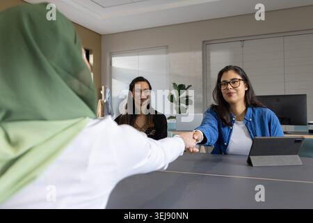 Diverse collaboratrici che stringono la mano a tavola in hijab e camicia con tablet e spazio di copia Foto Stock
