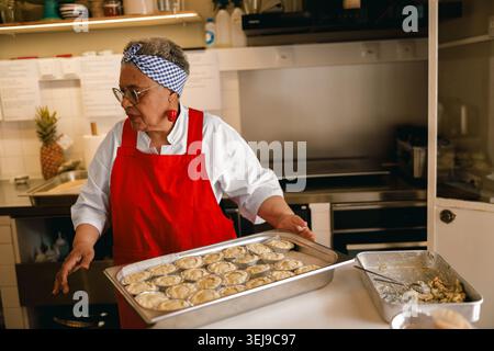 Chef donna Senior Black in grembiule rosso con vassoio di mini torte in un ambiente professionale Foto Stock